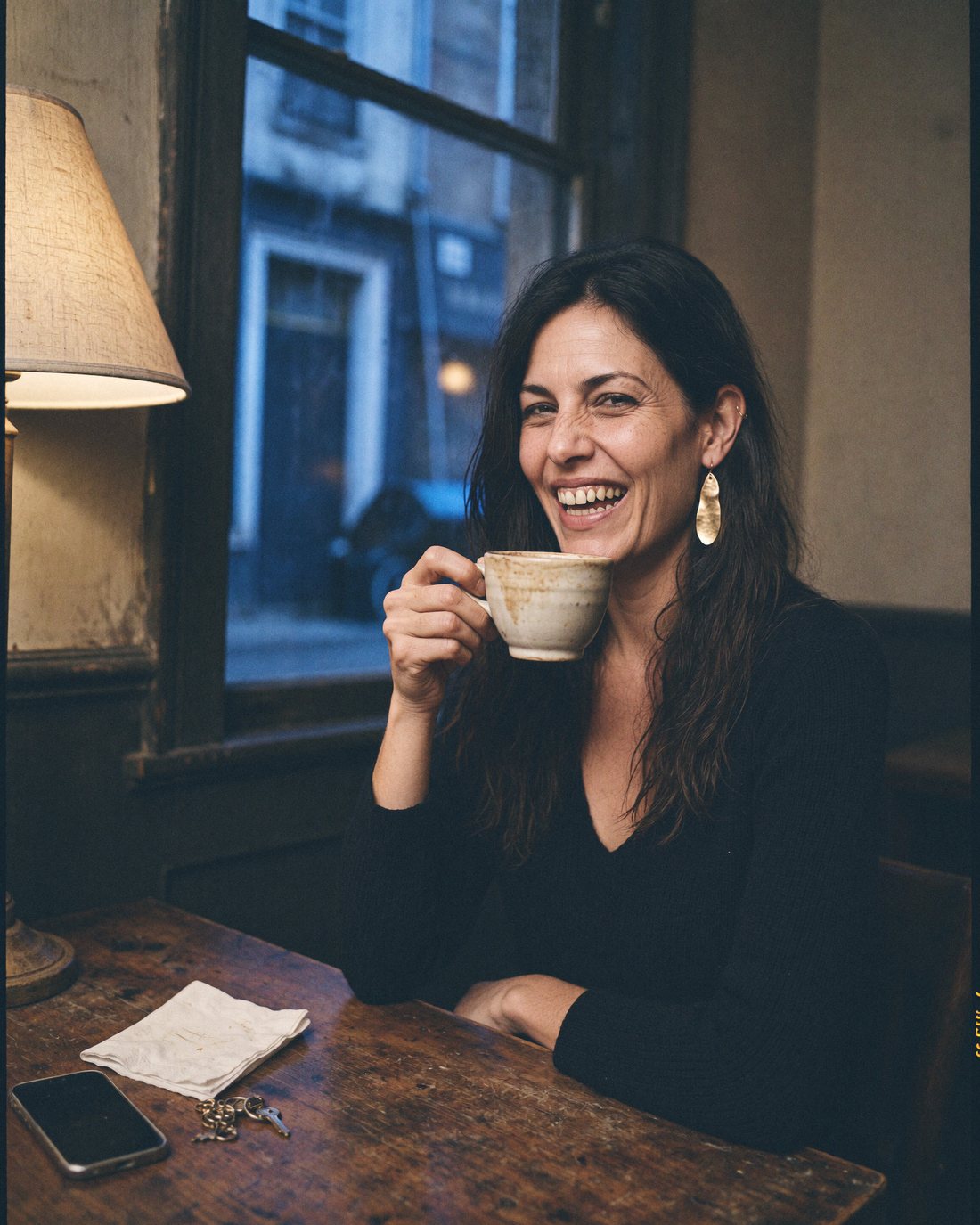 Lizzy Veyra laughing at a café table late in the evening, a cup in her hand.