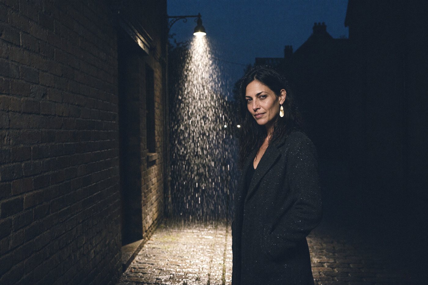 Lizzy Veyra standing under a street lamp in the rain on a narrow cobblestone street.