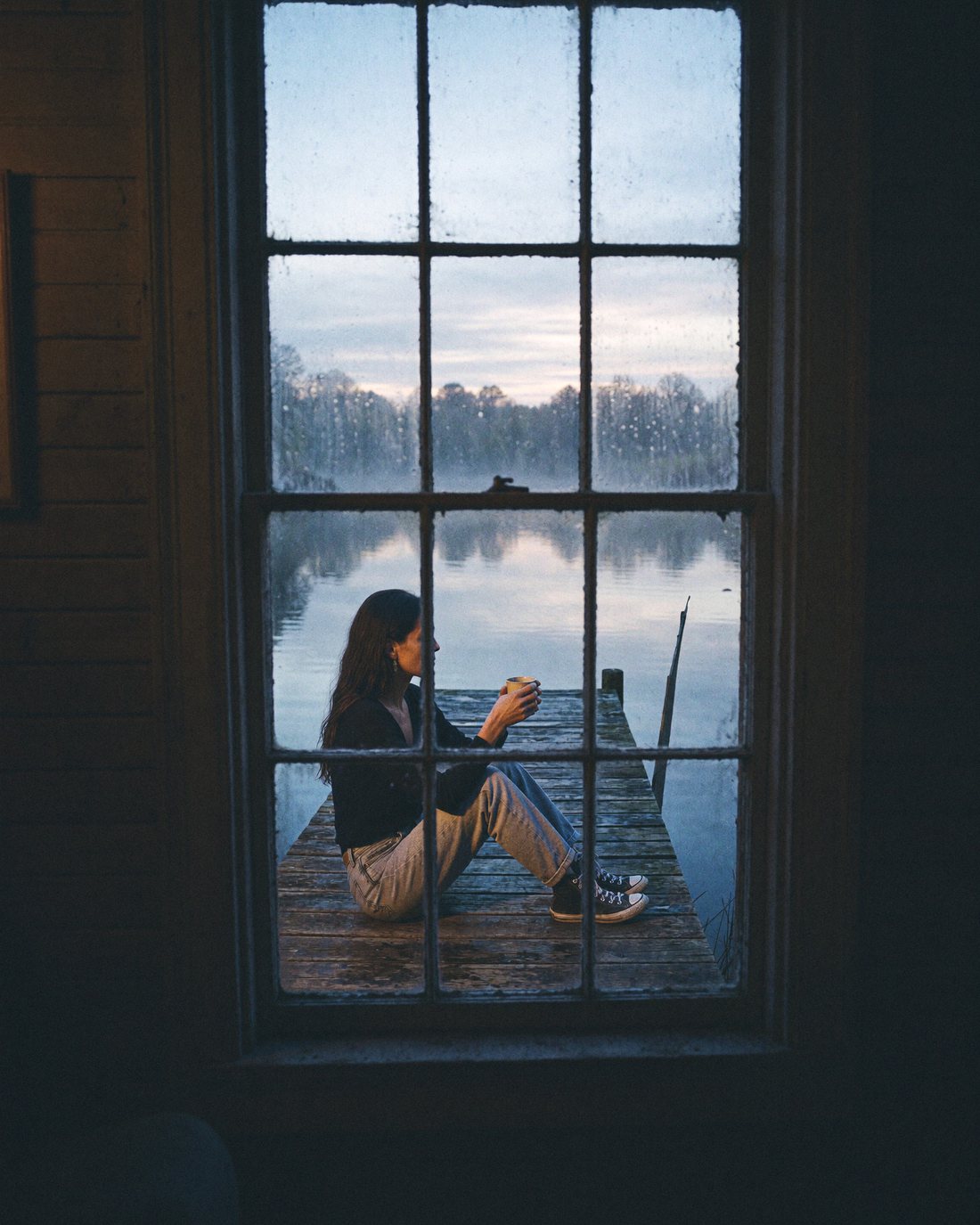 Seen through an old cabin window: Lizzy Veyra sitting at the end of a wooden dock in the early morning.