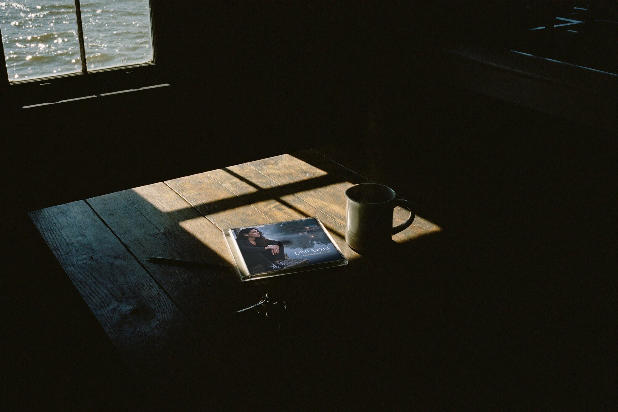 A CD of The River Knows My Name on an old wooden table in a stripe of morning sunlight, next to a cup of coffee.